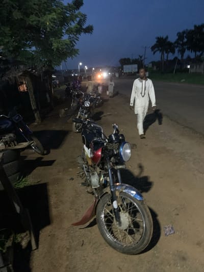 The image depicts a scene in Gbeleko, Nigeria, at dusk. A man wearing a white traditional garment stands on a dirt road adjacent to several parked motorcycles. A motorcycle in the foreground is the most prominent feature, showing wear and possible disrepair. Streetlights illuminate the background, where other motorcycles are parked. The environment includes trees and vegetation characteristic of the region. The sky is a dark blue, suggesting the early evening.