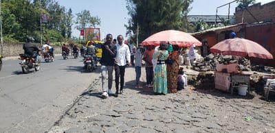 The image shows a busy street scene in Rubavu, Rwanda, located at latitude -1.668 and longitude 29.268.  The street is paved with uneven cobblestones. On the left side of the street, several motorbikes are moving in both directions.  A yellow taxi is parked at the curb. On the right side of the street, there are a few people congregating under a red umbrella.  One of the people appears to be selling something from a small cart.  A second red umbrella, slightly off to the right, is also being used to provide shade for a small street vendor.  Two men are walking toward the camera.  The overall scene is one of bustling activity, with people going about their daily business.  The weather is sunny and the sky is clear.