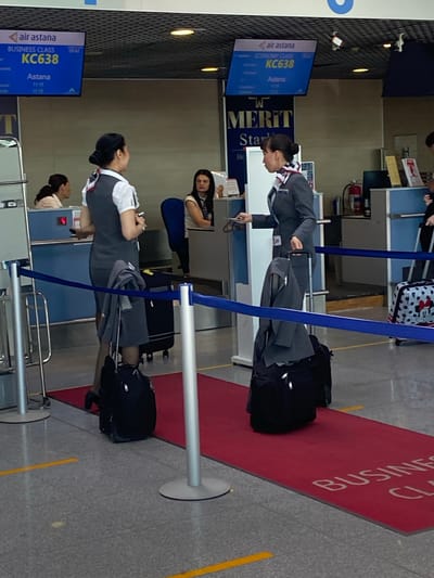 Two female flight attendants in grey uniforms and white shirts with patterned scarves stand at what appears to be an airport check-in or boarding area. They are positioned on a red carpet marked with "BUSINESS CLASS" lettering on the floor. Both women have their hair pulled back and are holding various items. The flight attendant on the left is facing the other, and the one on the right is looking towards the camera. In the background, an electronic display screen shows "AIR ASTANA" and flight information, including "KC638" and destinations. Other passengers and airport infrastructure, such as check-in counters and luggage carts, are visible. The overall scene depicts a typical airport environment during a flight departure or arrival. The presence of "AIR ASTANA" indicates a location where this airline operates, and the term "BUSINESS CLASS" suggests a premium service area. Given the context of an airport and an airline operating internationally, a specific city or country cannot be determined without further information.