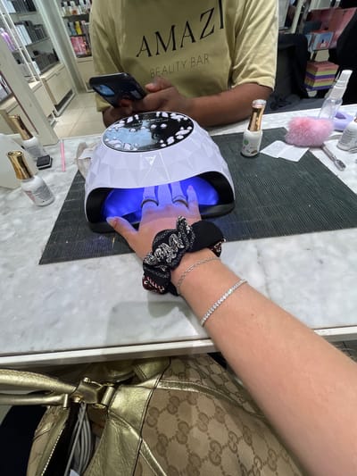 A person is having their nails done, placing their hand in a UV nail lamp for gel curing. A manicurist, wearing a tan t-shirt with "AMAZI BEAUTY BAR" printed on it, is operating the lamp and possibly taking a picture with their smartphone. The person getting their nails done is wearing a black and white patterned scrunchie and a tennis bracelet. A handbag is resting nearby. The setting appears to be a nail salon, based on the bottles of nail polish, tools, and shelving of products visible in the background. This is likely at Amazi Beauty Bar in Johannesburg, South Africa.