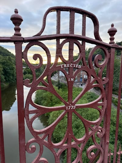 The image shows a close-up of a maroon, ornate iron bridge railing. The railing has decorative scrollwork and is embossed with the words "ERECTED IN 1779". In the background, through the gaps in the railing, a river flows on the left, and a town with buildings and a road can be seen on the right. Green trees and foliage line the riverbank and the hills behind the town. The sky above is overcast with hints of sunset peeking through the clouds. This is the Iron Bridge in Ironbridge, United Kingdom, famous as the world's first major iron bridge. There are no people or animals visible in the frame.