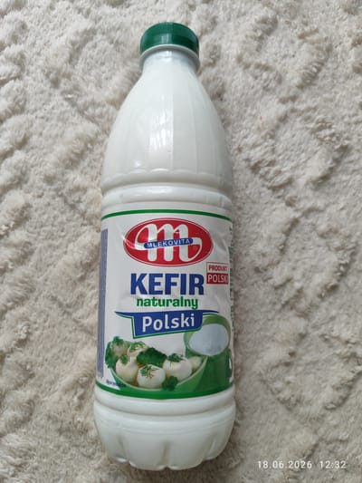 A white plastic bottle of Mlekovita Kefir Naturalny Polski is positioned upright on a white fluffy blanket. The bottle has a green cap. The label on the bottle is predominantly white with red and blue accents. At the top of the label, there is a red oval logo with the letter "M" in white. Below this, the text "MLEKOVITA" is visible.
The central part of the label features the word "KEFIR" in large blue letters, followed by "naturalny" in smaller green letters. Underneath "naturalny," the word "Polski" is written in large blue letters within a green banner. To the right of "KEFIR," a small red rectangle contains the text "PRODUKT POLSKI" in white letters.
At the bottom of the label, there is an illustration of a bowl containing small white balls, likely cheese, garnished with dill and broccoli. A green pitcher is also depicted. The text "Zdjęcie symboliczne" (Symbolic photo) is visible near the bottom edge of the illustration.
The bottom right corner of the image shows a timestamp: "18.06.2026 12:32".
The lighting appears to be artificial, and the scene is likely indoors. The overall impression is of a product shot, focusing on the kefir bottle. There are no people visible.