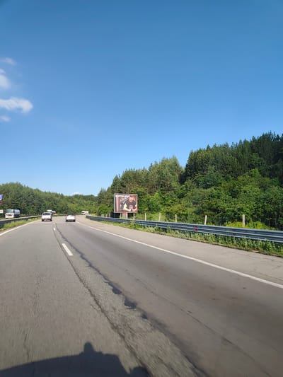 This image depicts a highway scene with a bright blue sky and lush green trees lining the roadside. Several vehicles, including cars and a truck, are traveling on the road. A billboard featuring a man with text above him is prominently visible on the right side of the road. The scene appears to be set in a natural, wooded area, likely outside of a densely populated urban center. The location is Potop, Bulgaria.
