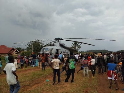 In Beni, Democratic Republic of the Congo, a large grey helicopter is landed on an unpaved, reddish-brown dirt clearing under a heavily overcast sky. The helicopter, featuring a prominent main rotor with five blades and a tail rotor, is surrounded by a crowd of approximately 50-70 individuals. The crowd consists of adults and numerous children, many of whom are observing the aircraft from various distances. Some individuals are positioned close to the helicopter's fuselage, particularly near an open side door, with a concentration of people seemingly interacting with the helicopter's interior. In the foreground, several young males stand on a dirt path, facing the helicopter. To the left background, two structures with distinct red-tiled roofs are visible, indicating a residential area. Beyond the helicopter, dense green foliage and trees form the horizon. A bicycle is present near the crowd on the right side of the frame.