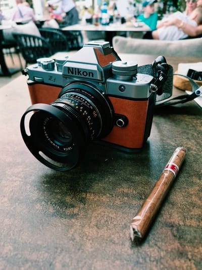 In Đenovići, Montenegro, a vintage Nikon ZF camera with a brown leather accent is positioned on a textured, dark brown table. Attached to the camera is a Leica Summicron 1:2/35 lens. In the foreground, a single cigar, wrapped in clear plastic and bearing a red and white band with gold lettering, lies to the right of the camera. The background is blurred, showing an outdoor seating area with people and tables, suggesting a casual, public setting like a cafe or restaurant.
