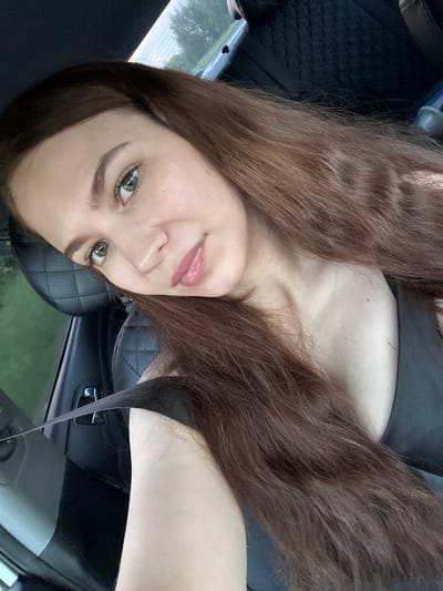 The image shows a young woman with long, brown hair, sitting in the driver's seat of a car. She is looking towards the camera with a slight smile. The interior of the car is visible, including a black leather seat and a seatbelt. The woman is wearing a black tank top. Outside the car, greenery and trees can be seen, suggesting a rural or suburban environment. The overall lighting suggests it is daytime. The location within the city of Кварса, Russia, cannot be determined from the image alone.