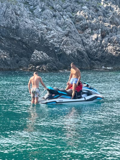 Two men are on a Yamaha jet ski in the clear, blue-green waters off the coast of Dubraljevina, Montenegro. The sun is shining brightly, illuminating the choppy water. One man, wearing blue patterned swim trunks, stands waist-deep in the water, holding onto the jet ski. He has a muscular build and appears to be enjoying the day. The other man, wearing red swim trunks and blue patterned shorts, sits on the back of the jet ski. He has a towel draped over his shoulders. The jet ski is white with blue accents and has "YAMAHA" written on the side. In the background, a steep, rocky cliff face rises from the water, covered in sparse vegetation. The time of day appears to be late morning or early afternoon, given the strong sunlight and the shadows cast by the rocks. The scene conveys a sense of leisure and adventure.