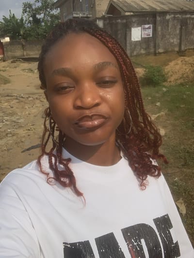 The image depicts an outdoor scene in the city of Anantigha, Nigeria. A female individual with dark complexion and reddish-brown braided hair occupies the foreground, facing the camera. She wears large silver hoop earrings and a white t-shirt with a distressed black graphic; the letters 'MADE' are partially visible on the shirt's front. Her lips appear glossy, and a sheen is present on her forehead and cheeks. The background consists of uneven bare earth and sparse vegetation, including trees and bushes. A concrete block wall extends horizontally, with two white documents affixed to its surface. Beyond the wall, a two-story building with a light-colored facade and a dark corrugated metal roof is visible, alongside another single-story structure featuring a similar roof. The sky overhead is partially cloudy.