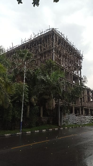 An unfinished building with scaffolding stands amidst dense greenery. The structure appears to be multi-storied, with an exposed framework. Some vegetation has begun to grow on the building itself. Adjacent to the building, a wet road surface reflects the overcast sky. The scene conveys a sense of abandonment and nature reclaiming an incomplete construction project.