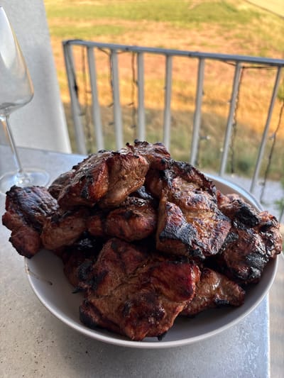 This is a close-up, high-angle shot of a white bowl filled with grilled meat. The meat appears to be small, bite-sized pieces, some of which are slightly charred, indicating they were cooked on a grill. The cuts of meat are varied in shape and size, with visible grill marks on several pieces. The bowl is positioned on a textured, metallic surface, possibly a table or countertop.

In the background, a metal railing is visible, suggesting an outdoor setting like a balcony or patio. Beyond the railing, a blurred landscape of dry, golden-brown grass and a hint of green vegetation is discernible. The lighting suggests it might be daytime, with the sun casting warm tones on the scene. A clear wine glass lies on its side in the lower left corner of the image, partially obscuring the view of the railing and the landscape. The overall impression is of a meal being prepared or served outdoors, likely in a casual setting. There is no visible text in the image.