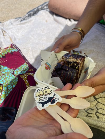 Two people are sharing a slice of chocolate cake outdoors. One person's arms are visible, holding the cake container and presenting it. Their wrist bears two bracelets, one gold and one silver. The other person's hand is holding several wooden spoons. The cake is dark brown, topped with chocolate drizzle and chopped nuts, and appears to be served on a paper liner within a white container. A colorful African print fabric is visible to the left of the cake. The ground appears to be sand or a light-colored blanket. The presence of a phone in the foreground suggests a casual setting, possibly a picnic. The specific location in Badalona, Spain, is not identifiable from the visual information provided.