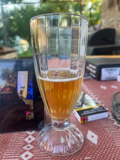 A close-up, eye-level shot captures a half-full glass of golden-hued beer with a frothy head, resting on a table. The glass has vertical fluting and a decorative, rippled base. The table is covered with a red and white patterned cloth. In the background, a black smartphone is visible, displaying an image with vibrant blue and purple accents, possibly a game interface. To the right of the beer glass, a pack of cigarettes sits on the table. The scene appears to be outdoors, suggested by the soft-focus bokeh of green foliage and sunlight in the upper background, indicating a daytime setting. The overall impression is one of casual relaxation. The location is identified as Kamenets, Bulgaria.