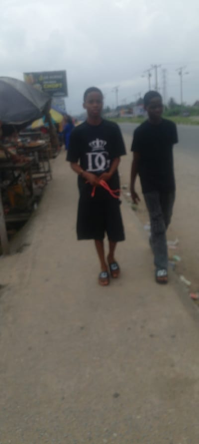 The image depicts two young males walking side-by-side along a paved pedestrian walkway adjacent to a road. The setting appears to be an outdoor market or roadside vendor area, possibly in Yenegoa, Nigeria, given the style of stalls and signage visible in the background. The sky is overcast and the lighting is dim.
The male on the left, closer to the foreground, is wearing a black t-shirt with a large white graphic of a crown above the letters "DQ" and a red string tied around his hands. He is also wearing black shorts and brown sandals.
The male on the right, slightly behind and to the right of the first male, is wearing a black t-shirt and dark pants. He is also wearing sandals. His left arm is bent at the elbow, and his hand is near his waist. Both individuals appear to be walking forward.
The background includes what looks like roadside stalls with various items displayed, a large billboard with text and graphics, and utility poles. The ground is a light-colored, rough pavement, with some scattered debris. The overall image quality is somewhat blurred, suggesting movement or a low-resolution camera.