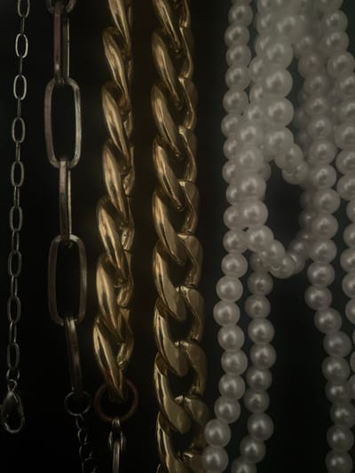 The image displays a close-up, vertical view of several strands of jewelry against a dark background. From left to right, there is a thin, silver-colored chain with an oval link pattern, a thicker, more robust silver-colored chain with larger, rectangular links, two thick, gold-colored curb chains with substantial, twisted links, and multiple strands of white pearl necklaces. The lighting is dim, creating shadows that highlight the textures and metallic sheen of the chains and the smooth, lustrous surface of the pearls. There are no people or identifiable landmarks visible. The image does not contain any information that would allow for the determination of its location in Torquay, United Kingdom.
