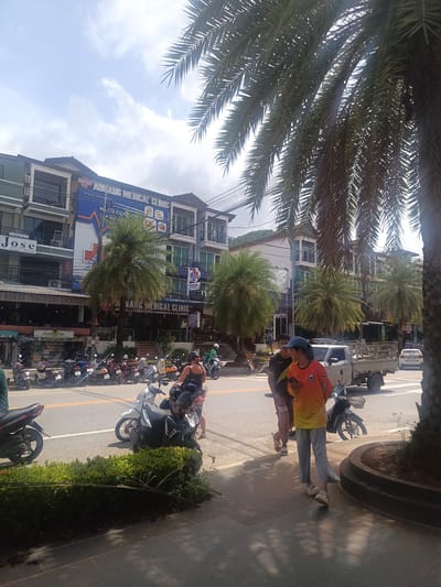 The image depicts a street scene in Ban Ao Nang, Thailand, with multiple businesses visible. On the left side, there is a restaurant named "Jose" and another establishment offering "BURGER THAI FOOD COCKTAILS". Further down, there are two "ADNAMANG MEDICAL CLINIC" signs and a "NANG MEDICAL CLINIC" sign. Several parked motorbikes line the street. In the foreground, two individuals are walking on the sidewalk. One wears an orange shirt and grey pants while holding a phone. A palm tree dominates the right side of the frame. A light-colored truck is on the road. The sky is partly cloudy.