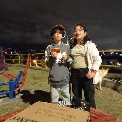 Two children stand outside at night. One child holds a slice of pizza, wearing a gray Adidas sweatshirt and brown hat. The other child stands next to them, wearing a light green shirt and a white jacket. A light-colored dog is standing in the background on the grass behind them, with city lights visible in the distance. There is a playground structure in the foreground and cardboard boxes to the right.