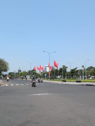 The image captures a sunny day on a wide avenue in Tuy Hòa, Vietnam. The view is along the street, showing the road stretching into the distance. Red Vietnamese flags are displayed along the median strip, attached to lampposts. Several motorbikes are traveling along the road. The sky is clear and blue. Green trees and buildings are visible alongside the road.