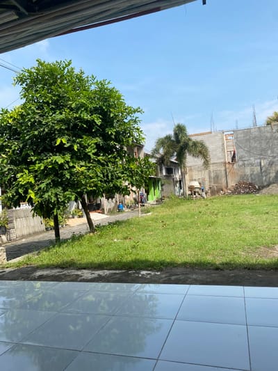 The image shows a view from a patio in Kecamatan Piyungan, Indonesia, with coordinates -7.827 latitude and 110.482 longitude. The patio is made of light grey tiles. There is a large green tree in the foreground and a small palm tree in the background. The scene is a mix of paved areas and grass with some foliage and a small concrete wall in the background.