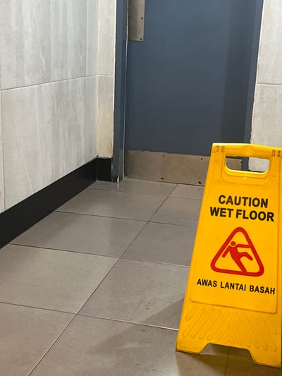 The image shows a yellow "Caution Wet Floor" sign, with the additional warning "Awas Lantai Basah" (Malay for "Beware Wet Floor"), placed on a tiled floor. The sign is located near a wall with a dark gray lower section and a light gray upper section. The tiles are gray, with a grid pattern. No people are visible. The location is likely in Bencongan Indah, Indonesia, although this cannot be confirmed forensically from the visual data.