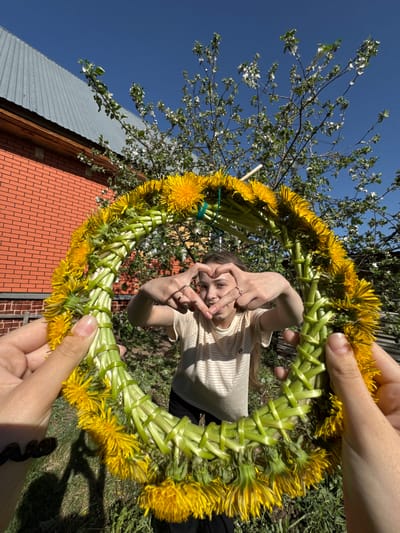 A person holds up a dandelion wreath in front of a young woman, who is looking through the wreath and making a heart shape with her hands. The wreath is made of green stems and yellow dandelion flowers. The person holding the wreath has fair skin. The young woman has long hair and is wearing a striped shirt. In the background, there is a tree with white blossoms, a blue sky, and a building with a gray roof and brick walls. The location is Кемуль, Russia.