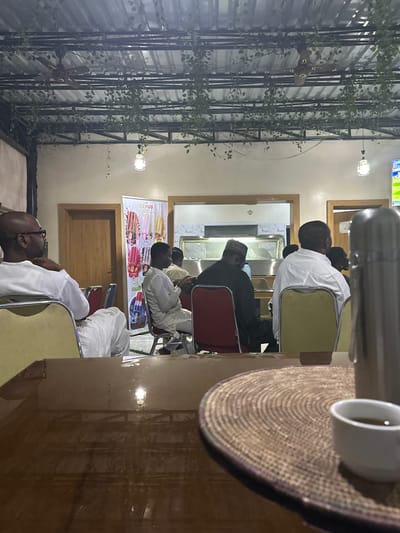 In Dandagoro, Nigeria, a group of individuals are seated in a casual indoor setting, possibly a restaurant or community hall. The scene is illuminated by artificial lighting and a ceiling fan is visible overhead, draped with artificial vines. In the foreground, a wooden table holds a woven placemat, a white teacup with a dark beverage, and a silver thermos. Behind the table, several people are seated in chairs facing away from the camera. One man in the foreground, on the left, is wearing a white tunic and glasses, looking to his left. A banner displaying colorful ice cream popsicles with the "ICEHUB" logo is visible on the left side of the frame. Further back, other individuals are seated, some wearing traditional attire, facing a serving area or presentation. The overall atmosphere appears relaxed and communal.