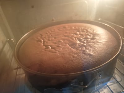 The media file shows a close-up, eye-level view of a chocolate cake baking in an oven. The cake is in a round, dark-colored springform pan, which is placed on the oven rack. The cake has risen significantly and has a bubbly, textured surface, indicating it is still in the process of baking. The interior of the oven is visible, with its walls illuminated by the oven light, creating a warm, bright glow that reflects off the cake. The image captures the moment of baking, suggesting a domestic setting, likely a home kitchen in Mińsk Mazowiecki, Poland. There is no visible text in the image. The overall mood is one of anticipation and domestic warmth, associated with the process of baking.