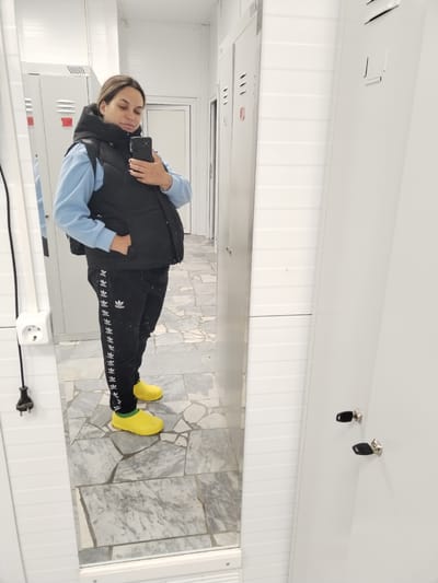 A woman is taking a mirror selfie in what appears to be a locker room. She is wearing a light blue hooded sweatshirt, a black puffer vest, black track pants with white Adidas stripes, and bright yellow rubber boots. Her hand is in the pocket of her vest. She is looking at her phone, which she is holding up to take the photo. The room has white paneled walls, grey and white marbled floor tiles, and several white lockers visible. A white electrical outlet is mounted on the wall to the left, with a black cord hanging from it. The background shows reflections in a large mirror. The image does not provide any specific location information that would indicate it is in the city of Foki, Russia.