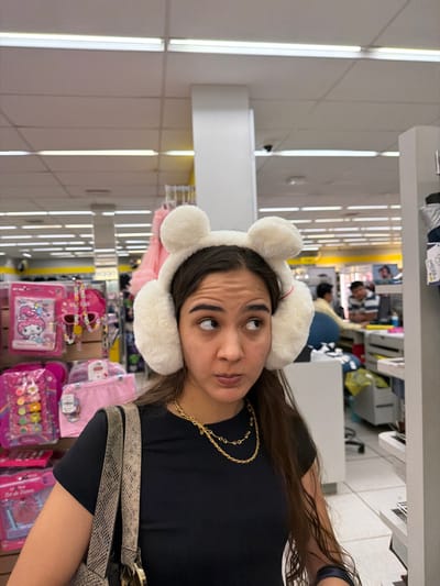 A young woman with dark hair and brown eyes poses in a store. She wears white fuzzy earmuffs with ears, a gold chain necklace, and a black shirt. A patterned purse strap is visible over her shoulder. Behind her is a store aisle filled with colorful merchandise, including pink stationery sets and children's items. The lighting is bright and fluorescent, typical of a retail environment.