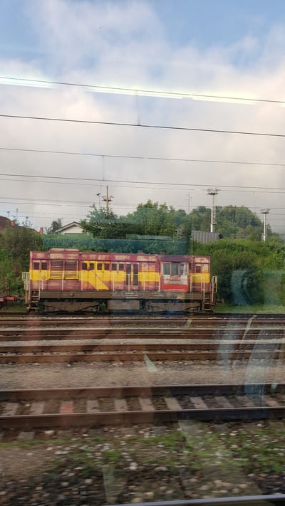 The image shows a weathered, yellow and red train engine parked on a set of railway tracks. The train has multiple compartments with windows and appears to be stationary. In the background, there are trees, bushes, and some buildings, including a house with a red roof. Overhead, there are power lines and utility poles. The sky is cloudy, with patches of blue visible. The scene is viewed through a reflective surface, likely a window, which causes some distortions and reflections. The ground around the tracks is covered with gravel and sparse vegetation. The overall impression is of an industrial or railway yard setting in Margecany, Slovakia.