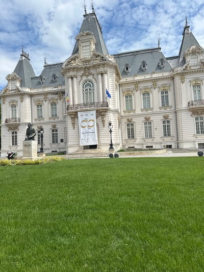 The image displays the Craiova Art Museum in Craiova, Romania. The museum is a grand, multi-storied white building with ornate architectural details, including many windows. A sign hangs above the entrance. In front of the museum is a green lawn. A statue of an individual stands to the left of the building's entrance. Two people are sitting near the base of the statue. The sky is visible in the background.