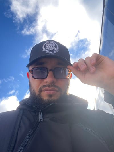 A man is taking a selfie outdoors. He is wearing a black baseball cap with a skull and barbell logo, sunglasses with tortoise shell frames, and a black jacket with zippers. He is holding the brim of his cap with his right hand, and the sun is shining brightly from the right, creating a strong backlight. The background shows a blue sky with white, puffy clouds. There is no discernible location in the city of Koskolovo, Russia, visible in the image.