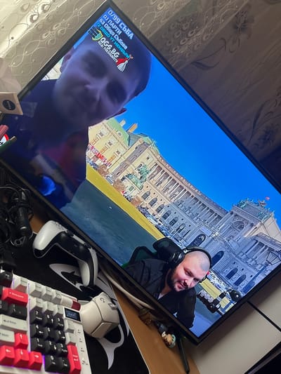 The image is a photograph taken indoors, focusing on a television screen displaying a live video feed. The video appears to be a livestream or broadcast featuring two men. One man, with a shaved head and wearing headphones, is positioned in the upper right portion of the screen, set against a backdrop of a grand, ornate building under a bright blue sky. The other man occupies the lower-left quadrant of the screen.

The foreground of the photograph shows a desk cluttered with various objects. A white PlayStation controller, a white Logitech mouse, and a keyboard with a mix of black, white, and red keycaps are visible. A figure toy sits on the desk near the TV. To the left of the desk are items, including a Nivea container, markers and other indiscernible materials. 

The text visible on the television screen, including "0/99 СЪБА", "63 Общо Събове", "DJ ПАРТИ", and "72 ОБЩО УТ МЕМБЪРИ" and the website "OGS.BG," suggests the video feed may be related to a gaming stream, online community, or entertainment broadcast in Bulgarian, while the building shown behind the one streamer is the Hofburg Imperial Palace in Vienna, Austria.