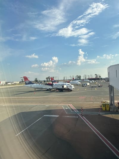 The image depicts an outdoor scene at an airport, likely Nikola Tesla Airport in Surčin, Serbia, given the presence of Air Serbia aircraft. Several Air Serbia airplanes are parked on the tarmac. One plane in the foreground, a turboprop aircraft, has "Air Serbia" and "TU-ALM" written on its fuselage. It is positioned on the tarmac, with its propellers visible. A service vehicle, possibly a fuel truck or baggage tractor, is parked next to it. In the background, more Air Serbia planes are visible, along with other airport infrastructure including a jet bridge or terminal building on the right. The tarmac is marked with various lines and symbols, indicating taxiways and parking positions. The sky above is blue with scattered white cumulus clouds. The overall scene is consistent with an operational airport environment under daylight conditions.