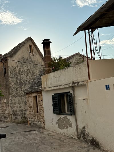 The image shows a narrow street in Primošten, Croatia, with two older stone buildings. The building on the left has a weathered stone facade and a grey slate roof with a prominent chimney. To the right, a lighter colored building features a single window with dark green shutters. Above this building, a corrugated metal roof structure is visible, supported by red metal poles. A blue street sign with the number "2" and text that appears to read "PUT NOVE" is affixed to the wall of the right building. The street is paved with cobblestones. The sky above is a pale blue with scattered white clouds. The scene is devoid of people or any obvious activity.