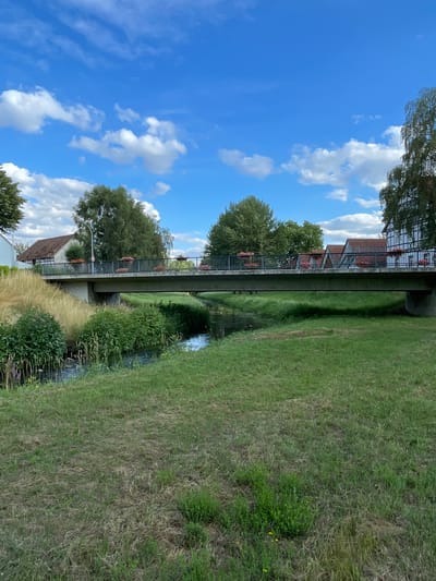The image shows a bridge over a small stream in Röllshausen, Germany. The bridge is made of concrete and has a metal railing. There are flowers planted in boxes along the railing. On either side of the stream are grassy banks with some trees and houses in the background. The sky is blue with white clouds.