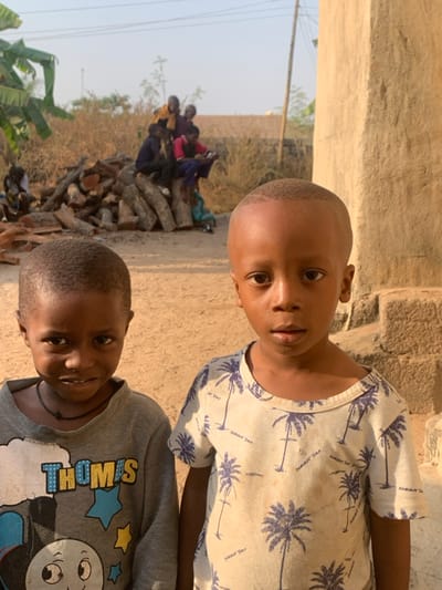 The image depicts an outdoor scene in Garaku, Nigeria, featuring two young Black males in the foreground. The child on the left, with dark skin and short dark hair, wears a grey long-sleeved shirt with a "Thomas" (Thomas the Tank Engine) graphic and a dark beaded necklace, exhibiting a slight smile towards the camera. The child on the right, with lighter brown skin and a short, faded haircut, wears a light-colored, short-sleeved shirt patterned with blue palm trees and "Summer Time" text, looking directly at the camera with a neutral expression.
In the midground, to the left of the foreground subjects, a large pile of cut logs or firewood is visible. Several other Black children are positioned on and around this woodpile, some seated, some standing, with their features indistinct due to distance; their clothing includes colors such as yellow, red, and blue. To the right in the midground, a section of a light-brown, textured building structure, possibly constructed from adobe or concrete, rises vertically. The ground beneath the subjects is bare dirt. The background comprises sparse, dry vegetation and a clear, bright sky, with faint overhead utility lines visible. The scene is illuminated by natural daylight.