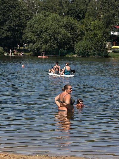 This media file captures a sunny day at a lake in Malakhovka, Russia, likely during summer. The scene is dominated by a large body of calm water with visible ripples reflecting the sunlight. The foreground features a sandy bank where several people are relaxing or preparing to enter the water.
The primary subjects are several individuals, mostly women, engaged in recreational activities on and around the lake. One woman in the foreground is seated on the bank, facing away from the camera, and appears to be looking out at the water. Behind her, several other people are in the water, some of whom are on stand-up paddleboards. One paddleboarder is partially submerged, with only their head visible above the water, looking towards the camera. Another individual is on a paddleboard further out on the lake. There are also people on the shore, some sitting and others standing. The overall atmosphere suggests a leisurely and enjoyable outing.
In the background, lush green trees and foliage line the bank of the lake, indicating a natural, park-like setting. A fence can be seen in the distance separating the treeline from the water. There are also some structures visible in the far background, including what appears to be a building with a red roof and a yellow vehicle, possibly part of a recreational facility or park.
The time of day appears to be midday, given the bright sunlight casting distinct shadows and illuminating the water. The weather is clearly warm and pleasant, as evidenced by the swimwear worn by the individuals and their presence in the water.
One of the paddleboards has visible text: "OCEAN". The emotions conveyed are primarily relaxed and happy, consistent with a day spent enjoying the outdoors and water activities.