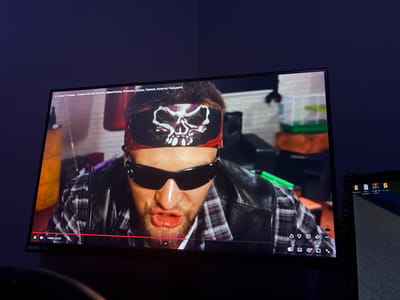 This is a still image from a YouTube video playing on a screen. The video features a man wearing a bandana with a skull design, sunglasses, a plaid shirt, and a black leather jacket. He has a beard and is making a facial expression with his mouth open, suggesting he is speaking or exclaiming. In the background, there are some objects that appear to be part of a room, including what looks like a white cage with a green and orange base, possibly for small animals.
The bottom of the screen displays YouTube video player controls, showing the title "6 Комнат! Обмани - Получи 500.000! (Бустер, ФрамеТамер, Кокошка, Кореш, Горилла, Куертов, Парадеич)". The current playback time is 1:28:08 out of a total of 23:27. There is also text indicating "NVIDIA G-SYNC". To the right of the screen, a laptop is visible, displaying desktop icons including "WORAR ZIP archive". The overall scene is dimly lit, suggesting an indoor setting, possibly a room or studio. The man's expression conveys a sense of surprise or strong emotion.
