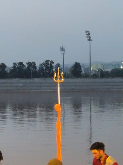 The image captures a scene at the Tawi River in Jammu, India, during dusk or dawn. In the foreground, a golden trident, a religious symbol, stands tall, draped with an orange cloth. The river's surface reflects the muted sky and the surrounding environment. In the background, a concrete embankment lines the river, beyond which are trees and distant buildings. Two tall stadium lights are visible against the sky. On the right side of the frame, a young man with dark hair and a beard is partially visible, wearing a red shirt and a patterned vest. He appears to be looking down.
