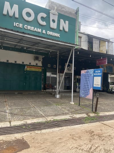 The image shows the exterior of two storefronts located at the coordinates -7.022, 107.676 in Baleendah, Indonesia. The storefront on the left is called "MOCON ICE CREAM & DRINK", with a green awning and a sign depicting a large cartoon glass of iced beverage. The storefront on the right is called "SALON MUSLIMAH" and appears to be a hair salon. The salon has a sign that lists various services, including "Creambath," "Smoothing," and "Hair Color." Both storefronts have chain-link fences extending from the sidewalk to the building. The ground in front of the stores appears to be a paved area consisting of small, irregularly shaped stones.  The background of the image shows the sides of two buildings, partially obscured by the awning and the second floor of a taller building. The weather appears to be cloudy.