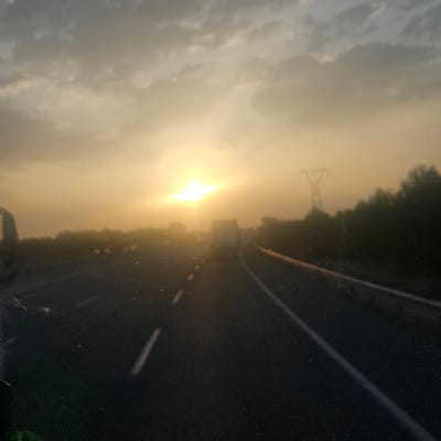 The image depicts a scene from a highway in Banyeres del Penedès, Spain, at either sunrise or sunset. The sun is low on the horizon, casting a warm, golden light across the sky and illuminating the clouds with hues of orange and yellow. The highway stretches out into the distance, with lane markings visible on the asphalt. On the right side of the road, there is a row of trees silhouetted against the bright sky, and a power line pylon can be seen further in the background. Several vehicles, including what appears to be a truck, are driving on the highway, their forms blurred by the low light and possibly motion. The overall impression is one of a serene journey through the Spanish countryside during the transitional hours of the day.