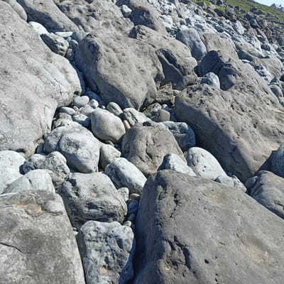 This is a close-up, eye-level, long shot of a rocky shore in Fanore, Ireland, on a sunny day. The scene is dominated by large, weathered grey rocks of various sizes, piled upon each other. Some rocks are smooth and rounded, while others are rough and irregular, with visible pores and markings. The texture of the rocks suggests they have been shaped by the elements over time. In the background, a grassy slope rises, with a few buildings visible at the top, indicating human habitation in the vicinity. The overall impression is one of a rugged, natural landscape. There are no people or animals visible in the image.