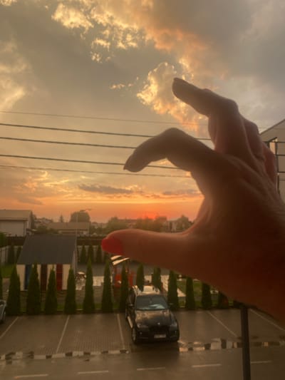 The image, taken in Novi Petrivtsi, Ukraine, captures a sunset scene with a human hand in the foreground. The hand is positioned with fingers extended, obscuring part of the view. The sky is filled with dramatic clouds illuminated by the setting sun, casting an orange glow. Below the sky, a landscape with houses and trees is visible, along with a parking lot containing a black SUV and a light-colored car. The ground in the parking lot appears wet. The overall impression is of a casual moment captured during twilight, with the hand adding a subjective element to the composition.