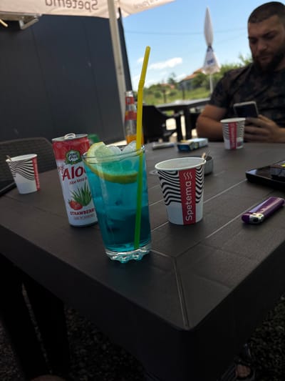 Here is a description of the image:

The scene is captured outdoors, on a dark-colored, textured table at what seems to be a restaurant or cafe. A man in a camouflage shirt sits partially in the frame, with his face blurred and his hand holding a smartphone.

The table is covered with several items: a lighter, two small paper cups with "Spetama" printed on them, a tall glass of blue liquid with ice and a lemon slice, a yellow straw, and a can of "PURE PLUS my Alo Aloe Vera Drink" with a strawberry flavor. The angle of the table suggests a close-up shot, focusing on the items on the table.

In the background, there are trees, a building, and the sky, which is partially visible and shows a clear, sunny day. The angle of the shot suggests a perspective from a table positioned somewhat to the side and slightly below the man.

It can be assumed this image was taken in Dolni Tsibar, Bulgaria.