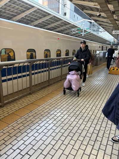 The image shows a man and a child standing near a bullet train at Tokyo Station in Chiyoda City, Japan (latitude: 35.682, longitude: 139.768). The man is wearing a black jacket and jeans and is holding a stroller with a child inside. The child is wearing a pink jacket. The train is white with blue and silver stripes. The platform is made of brick. There is a metal handrail next to the train. The man and child are standing on the platform facing the train.