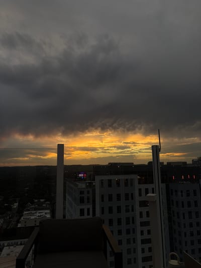 View from a high vantage point, likely a balcony or rooftop in the city of Austin, Texas, USA. The perspective is looking toward a cityscape, partially obscured by a dramatic sky with a strong contrast between dark storm clouds and a golden, sunlit horizon. Buildings of varying heights are visible, including residential and commercial structures. The sky is dominated by dark grey storm clouds on one side, and a bright, golden sunset or sunrise on the other. The built environment includes densely packed residential buildings, smaller structures along the ground, and green spaces. A portion of balcony furniture (chair or sofa) is visible in the foreground.