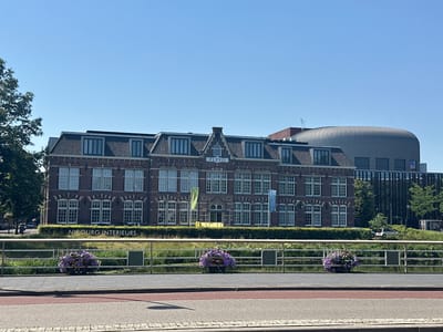This image captures a bright, sunny day in Zwolle, Netherlands. The scene features a prominent brick building with a dark tiled roof and numerous windows. The building has a sign that reads "FLEVO" above the entrance, and another sign on the lower part of the building facing the water that says "NIBOURG INTERIEURS". In front of the building, there is a canal with lush green foliage along its banks. A metal railing runs along the edge of the water, with two hanging planters filled with purple flowers adorning it. To the right of the brick building, a modern, curved, silver structure is visible, contrasting with the older architecture. The sky is clear and blue, indicating good weather. The presence of trees suggests an outdoor, urban or semi-urban setting. There are no people visible in the frame, and the overall mood is serene and picturesque.