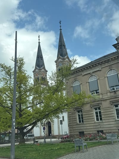 The image depicts the Église Saint-Symphorien (Saint Symphorien Church) in Illkirch-Graffenstaden, France, viewed from a low angle. The church features two prominent spires adorned with decorative crosses at their peaks, which rise into a cloudy sky. The church's facade includes clock faces and architectural details. Trees with light green foliage partially obscure the view of the church. To the right of the church, a section of a building is visible with arched windows, gray blinds, and classical architectural elements. In front of the building are flowerbeds and metal benches on a paved walkway and lawn. A gray pole with attachments stands in the foreground.