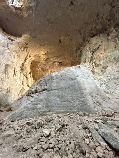 The image captures the interior of a cave in Karlukovo, Bulgaria, likely the Prohodna Cave, also known as the "Eyes of God" cave. A large, irregularly shaped boulder dominates the foreground, appearing to be made of grey rock. The boulder's surface is textured with various pits and indentations. Surrounding the boulder is a dense collection of smaller rocks and debris, suggesting a natural, undeveloped environment within the cave. The cave walls rise around the boulder, exhibiting rough, uneven textures. Light streams in from an opening higher up in the cave, illuminating parts of the walls and creating shadows. The overall impression is of a natural, geological formation, with the boulder being a significant feature within the cavernous space.