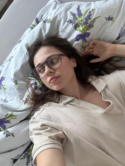 A young woman wearing glasses lies on her back on a bed with a white floral patterned sheet. She is wearing a beige shirt. Her left hand is raised. The image may have been taken in Kőröshegy, Hungary, but there are no elements in the picture that could confirm it.